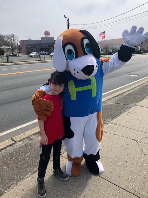 a person in a dog costume hugging boy in red shirt on sidewalk